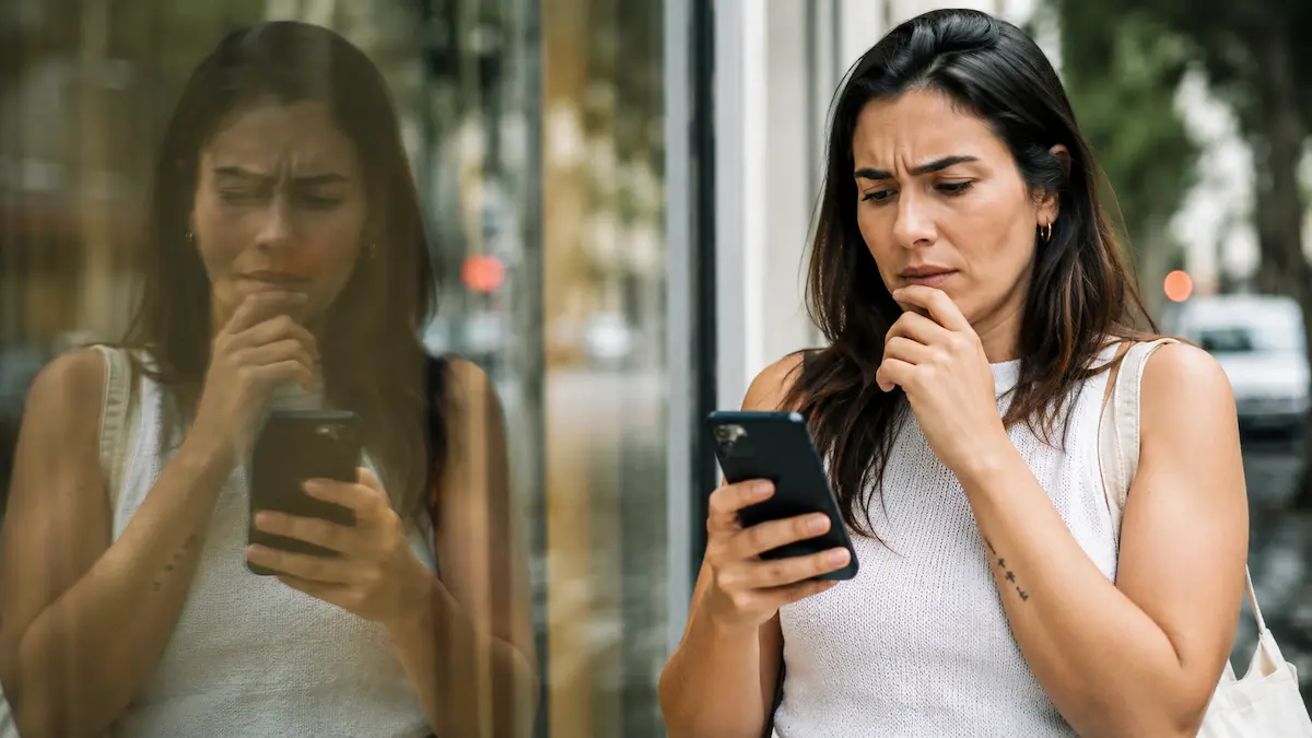 Pessoa olhando o próprio extrato bancário no celular com expressão de dúvida, refletida em uma vitrine